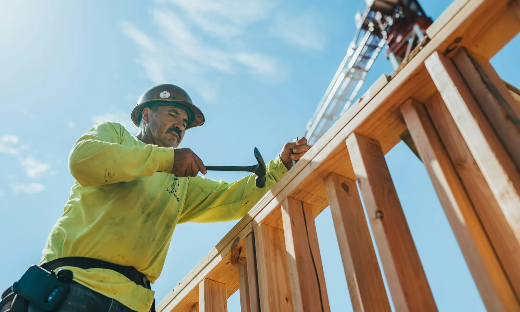 Contractor in a hard hat and work belt hammering a wood frame wall against a blue sky