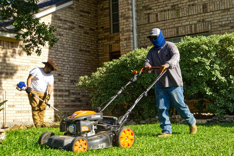 Two-man landscaping crew mowing and trimming a front lawn outside a brick house on a sunny day