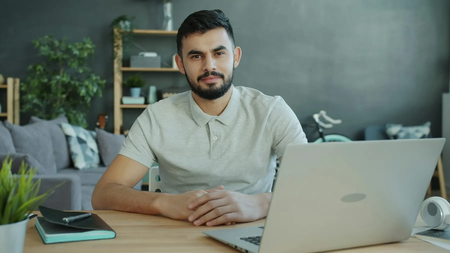 Bearded man in a gray polo sitting at a wooden desk with a laptop in a bright living room