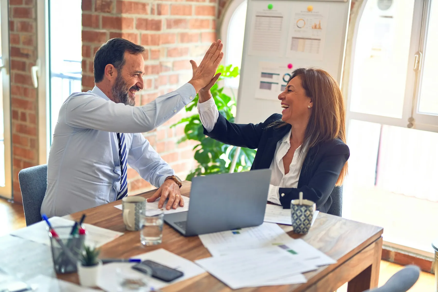 Two business partners high-fiving across a desk with paperwork