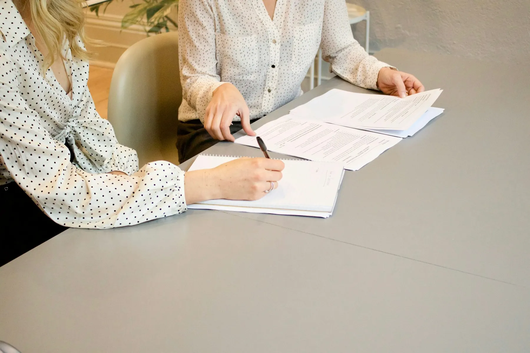 Woman signing a document at a gray table while a colleague points to the next page